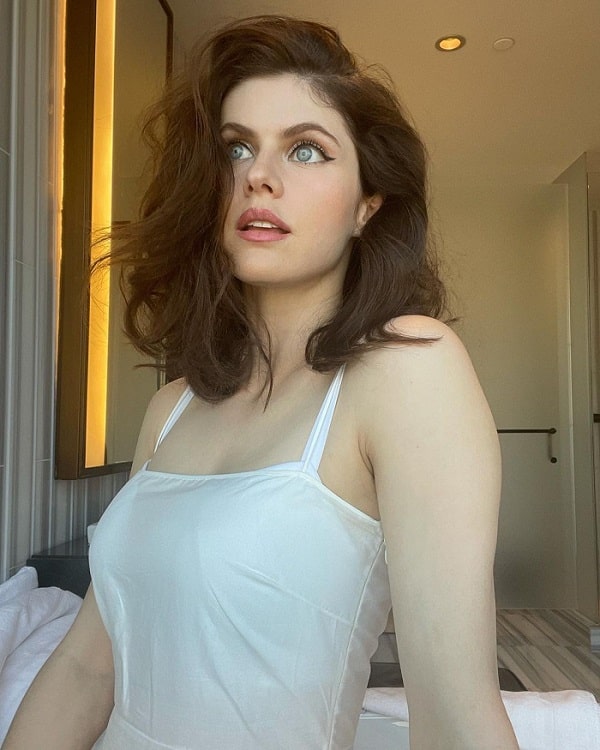 Alexandra Daddario poses in bed in a strappy white top.