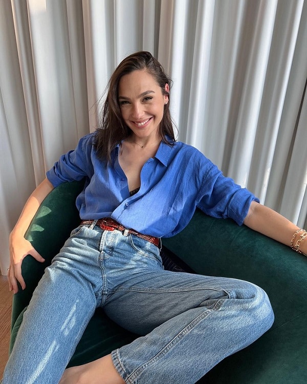 Gal Gadot poses on a sofa in tight jeans and a blue button-down shirt.