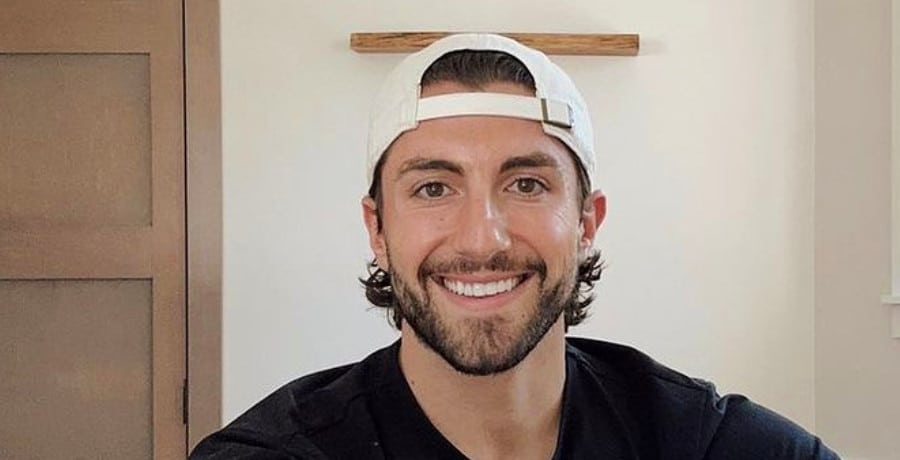 A man with brown hair and a beard wearing a white baseball hat backwards.