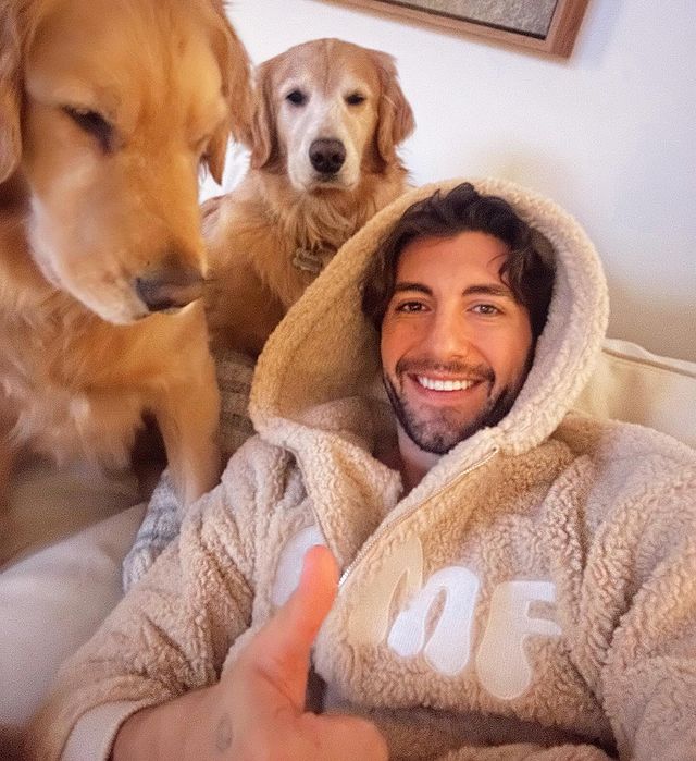 A man in an oversized hoodie with two golden retrievers next to him.