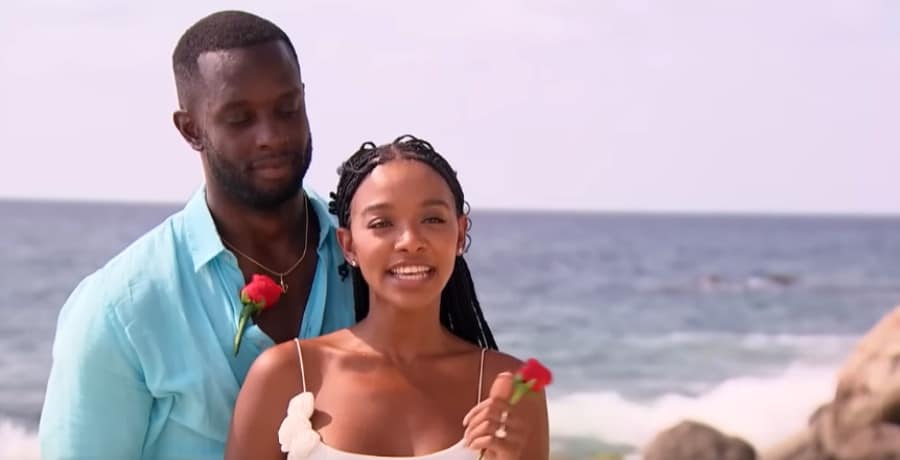 A Black man and a Black woman holding red roses on a beach.