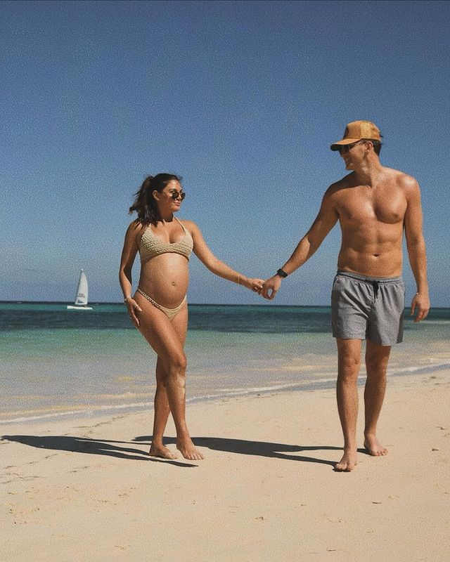 A pregnant woman and a man holding hands and walking on the beach. 